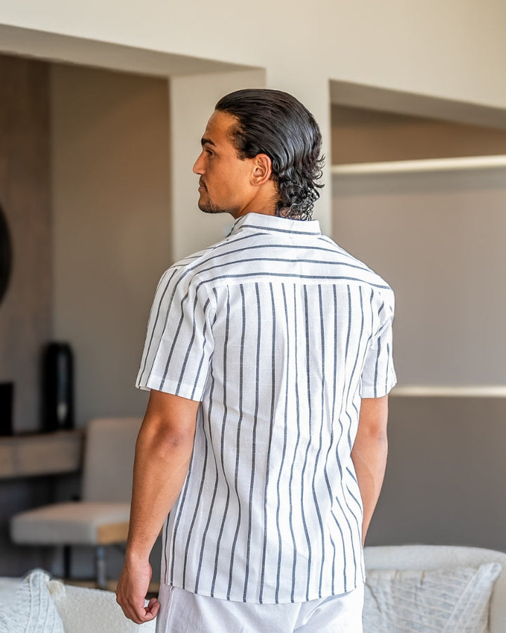 Homme portant la chemise d’été moderne Lyam en lin blanc à rayures foncées, vue de dos dans un intérieur élégant, avec manches courtes et coupe légère idéale pour un look estival raffiné.