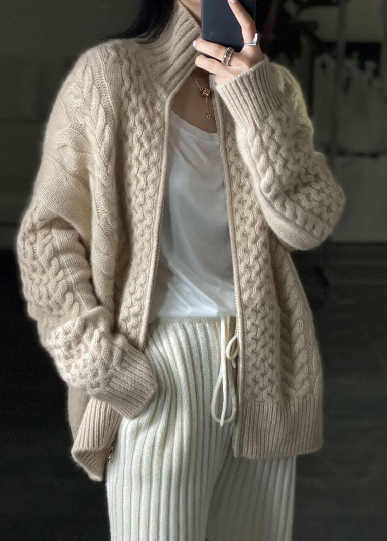 Femme portant un cardigan épais à col montant en maille torsadée beige, ouvert sur un t-shirt blanc, associé à un pantalon côtelé clair pour un style confortable et tendance.