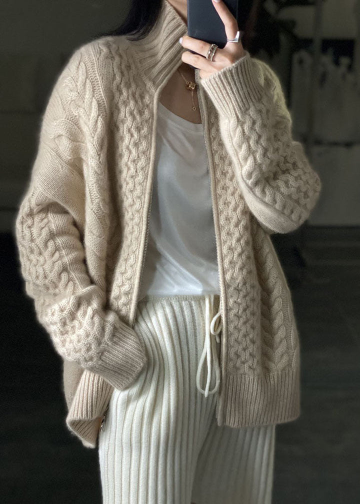 Femme portant un cardigan épais à col montant en maille torsadée beige, ouvert sur un t-shirt blanc, associé à un pantalon côtelé clair pour un style confortable et tendance.
