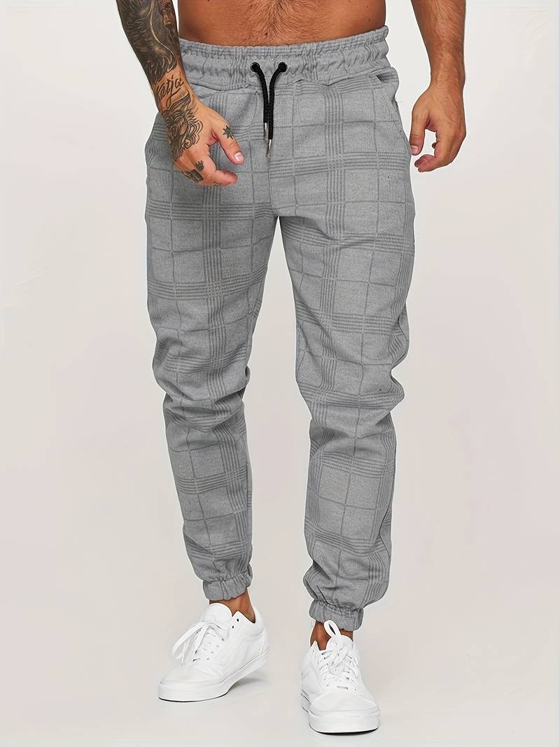 Pantalon de jogging pour homme de couleur grise avec motif à carreaux discrets, coupe moderne et ajustée, taille élastique avec cordon de serrage noir, chevilles resserrées, porté avec des baskets blanches, vue de face du bas du torse jusqu’aux pieds sur fond clair.