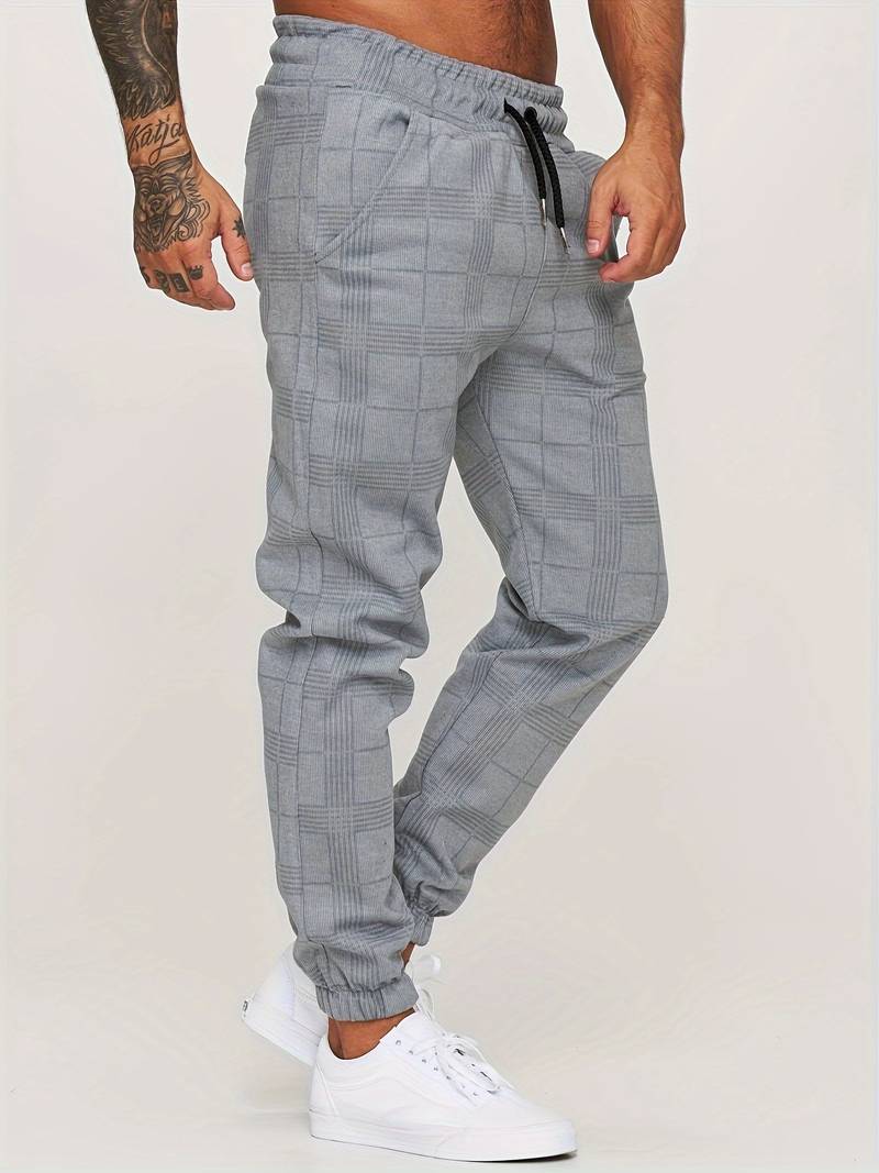 Pantalon de jogging pour homme gris à motif à carreaux discrets, coupe moderne et ajustée, taille élastique avec cordon de serrage noir, poches latérales, chevilles resserrées, vue de profil, porté avec des baskets blanches sur fond clair.
