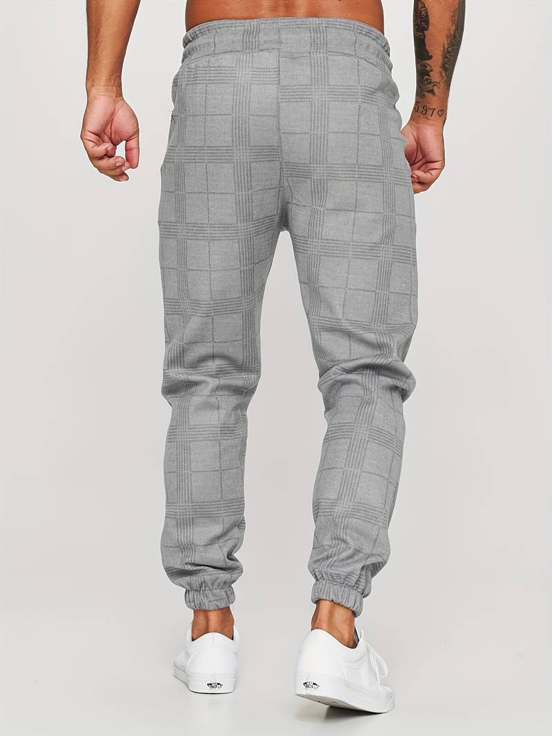 Pantalon de jogging pour homme gris avec motif à carreaux discrets, coupe moderne et ajustée, taille élastique, chevilles resserrées, vue arrière du bas du dos jusqu’aux pieds, porté avec des baskets blanches sur fond clair.