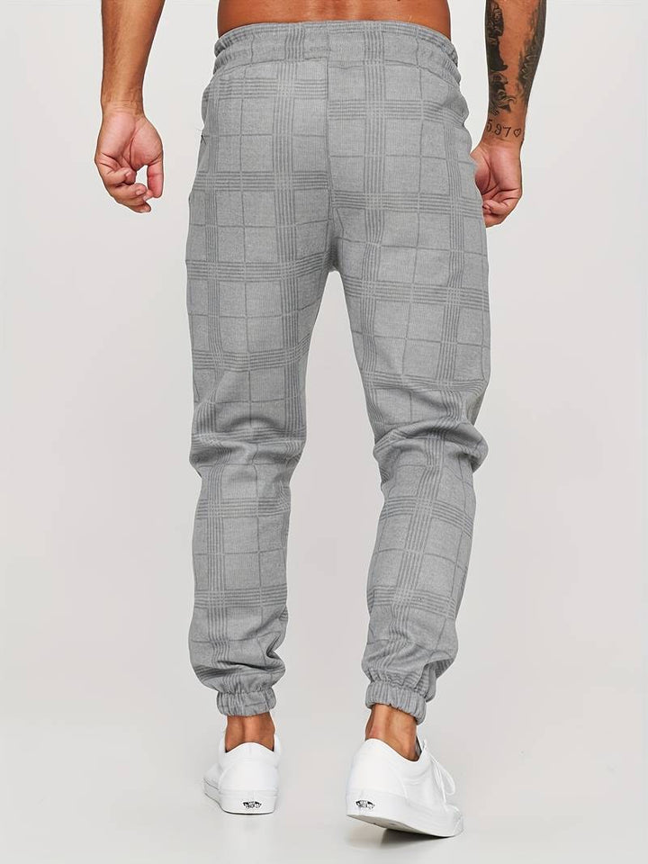 Pantalon de jogging pour homme gris avec motif à carreaux discrets, coupe moderne et ajustée, taille élastique, chevilles resserrées, vue arrière du bas du dos jusqu’aux pieds, porté avec des baskets blanches sur fond clair.