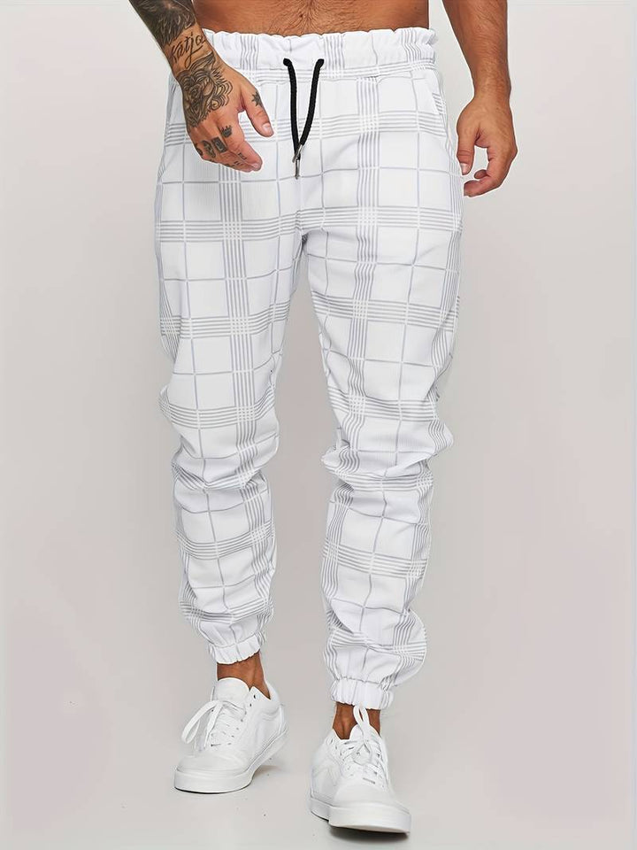 Pantalon de jogging pour homme blanc avec motif à carreaux discrets, coupe moderne et ajustée, taille élastique avec cordon de serrage noir, poches latérales, chevilles resserrées, vue de face du bas du torse jusqu’aux pieds, porté avec des baskets blanches sur fond clair.