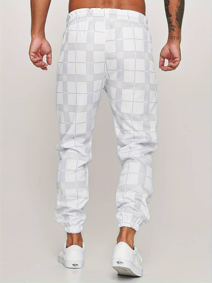 Pantalon de jogging pour homme blanc avec motif à carreaux discrets, coupe moderne et ajustée, taille élastique, chevilles resserrées, vue arrière du bas du dos jusqu’aux pieds, porté avec des baskets blanches sur fond clair.