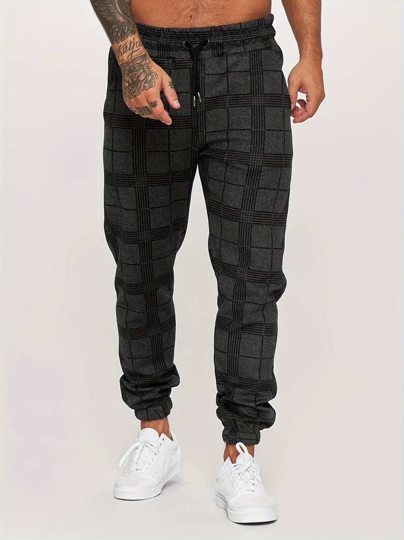 Pantalon de jogging pour homme noir à motif à carreaux discrets, coupe moderne et ajustée, taille élastique avec cordon de serrage noir, poches latérales, chevilles resserrées, vue de face du bas du torse jusqu’aux pieds, porté avec des baskets blanches sur fond clair.