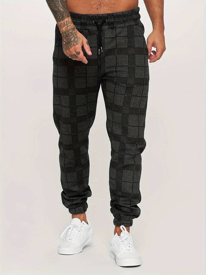 Pantalon de jogging pour homme noir à motif à carreaux discrets, coupe moderne et ajustée, taille élastique avec cordon de serrage noir, poches latérales, chevilles resserrées, vue de face du bas du torse jusqu’aux pieds, porté avec des baskets blanches sur fond clair.