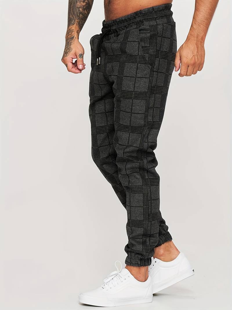 Pantalon de jogging pour homme noir à motif à carreaux discrets, coupe moderne et ajustée, taille élastique avec cordon de serrage noir, poches latérales, chevilles resserrées, vue de profil, porté avec des baskets blanches sur fond clair.