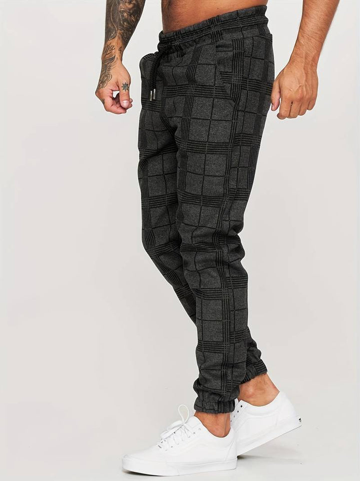 Pantalon de jogging pour homme noir à motif à carreaux discrets, coupe moderne et ajustée, taille élastique avec cordon de serrage noir, poches latérales, chevilles resserrées, vue de profil, porté avec des baskets blanches sur fond clair.