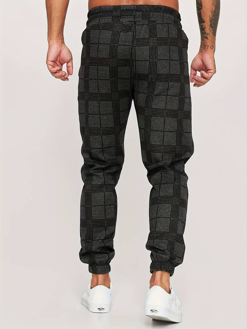 Pantalon de jogging pour homme noir à motif à carreaux discrets, coupe moderne et ajustée, taille élastique, chevilles resserrées, vue arrière du bas du dos jusqu’aux pieds, porté avec des baskets blanches sur fond clair.