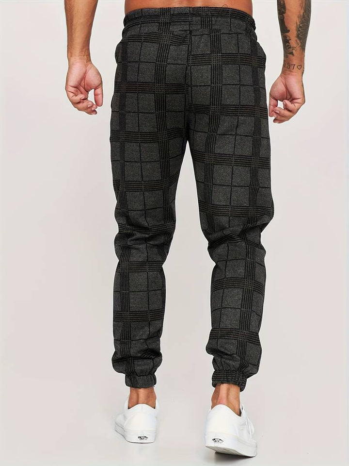 Pantalon de jogging pour homme noir à motif à carreaux discrets, coupe moderne et ajustée, taille élastique, chevilles resserrées, vue arrière du bas du dos jusqu’aux pieds, porté avec des baskets blanches sur fond clair.