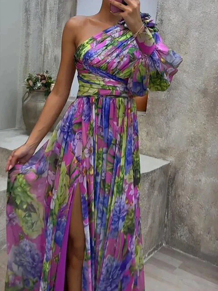 Une femme portant une robe longue asymétrique florale, vibrante et colorée, avec une fente haute sur la cuisse, mettant en valeur un mélange de fleurs violettes, vertes et bleues, affichant un style élégant et chic, parfait pour les événements formels ou les occasions estivales.