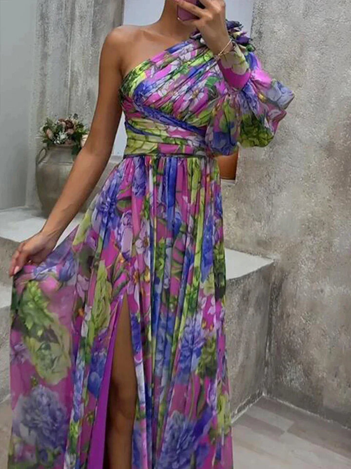 Une femme portant une robe longue asymétrique florale, vibrante et colorée, avec une fente haute sur la cuisse, mettant en valeur un mélange de fleurs violettes, vertes et bleues, affichant un style élégant et chic, parfait pour les événements formels ou les occasions estivales.