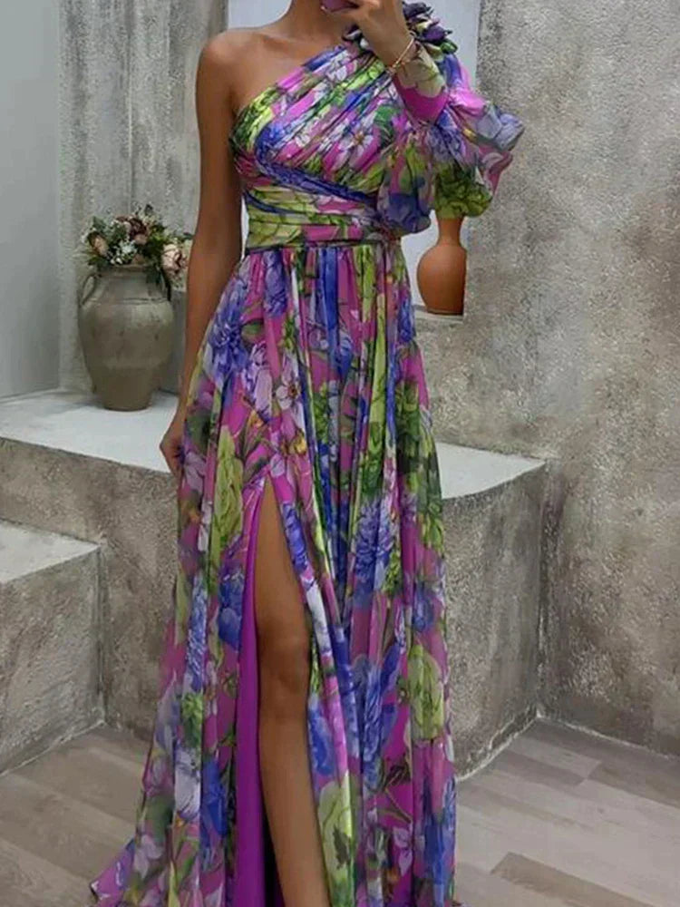 Une femme portant une robe longue asymétrique florale et colorée, avec une fente haute sur la cuisse, arborant des teintes vibrantes de violet, vert et bleu, dégageant un look chic et élégant, parfait pour les occasions formelles ou un événement estival.