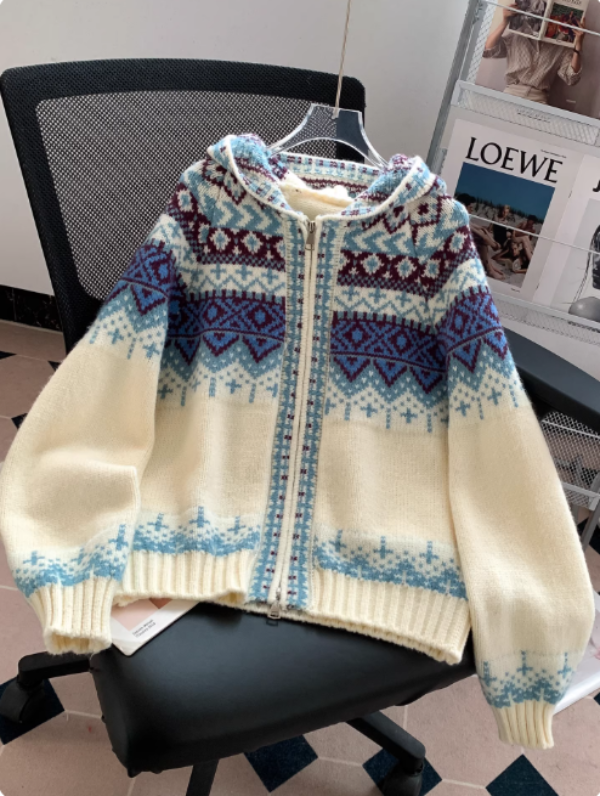 Un sweat à capuche blanc en maille zippée, orné de motifs géométriques bleus et bordeaux, est posé sur une chaise de bureau dans un intérieur moderne avec un porte-revues en arrière-plan.