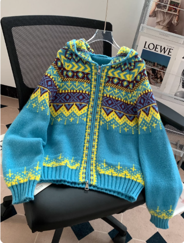 Pull à capuche en maille bleu turquoise avec fermeture éclair, orné de motifs géométriques jaunes, violets et bordeaux, posé sur une chaise de bureau dans un intérieur moderne avec un porte-magazines en arrière-plan.