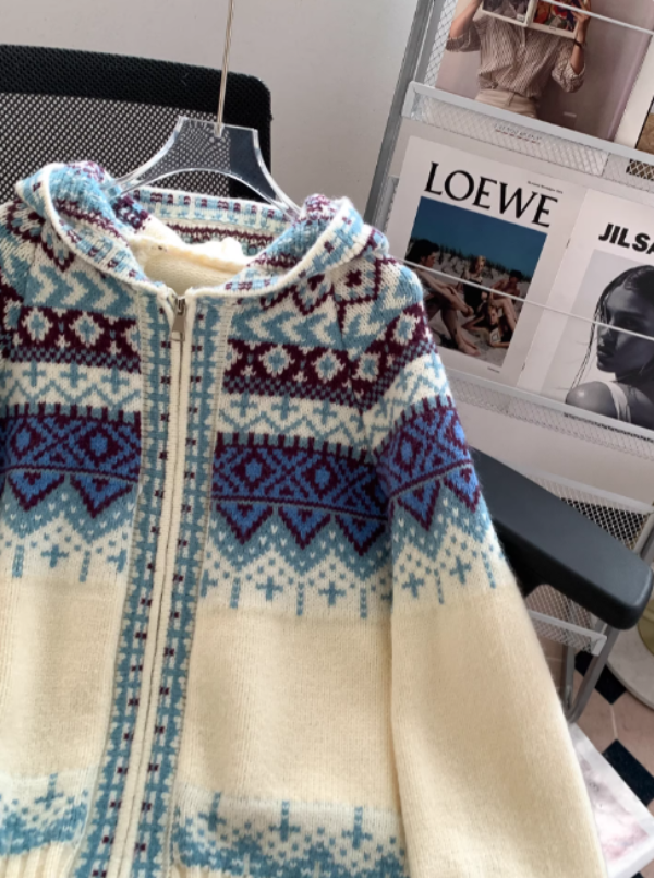 Gros plan sur un sweat à capuche blanc en maille zippée, orné de motifs géométriques bleus et bordeaux, présenté sur une chaise de bureau dans un intérieur moderne avec un porte-revues en arrière-plan.