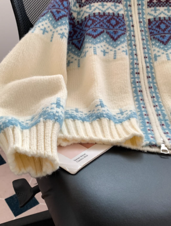 Gros plan sur la manche et le bas d'un pull à capuche blanc en maille, orné de motifs géométriques bleus et bordeaux, avec des poignets côtelés, posé sur une chaise de bureau avec un magazine en dessous.