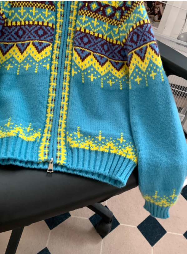 Gros plan sur le bas et la manche d’un pull à capuche en maille bleu turquoise, avec fermeture éclair et motifs géométriques jaunes, violets et bordeaux, posé sur une chaise de bureau.