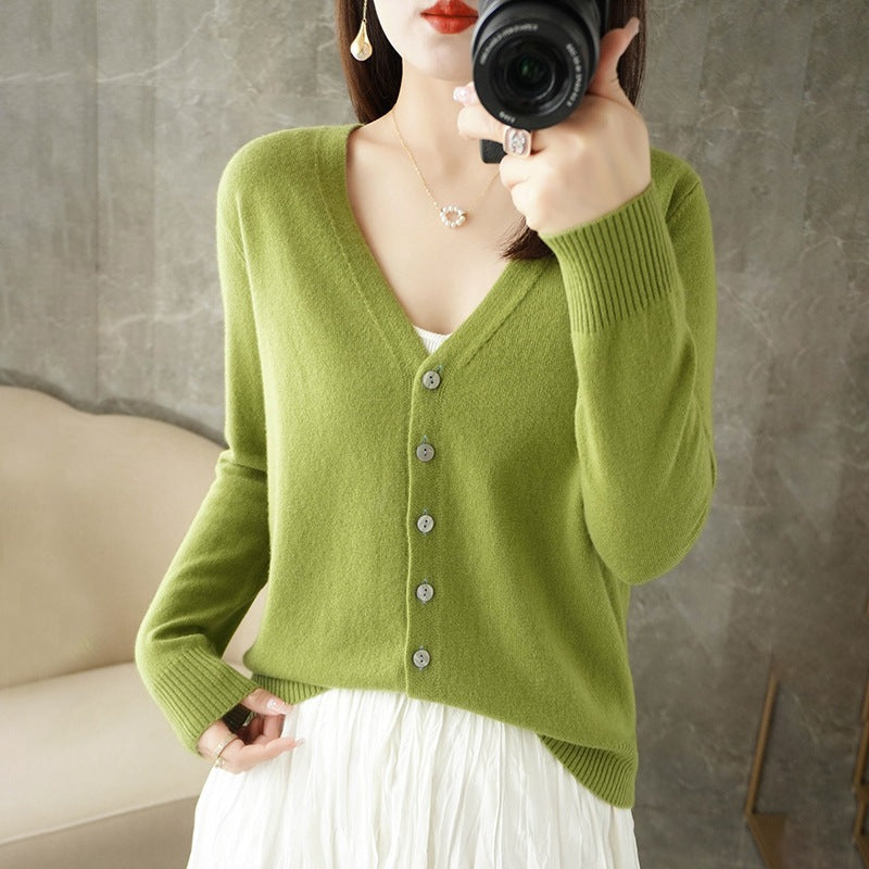 Femme portant un cardigan vert olive à col en V, doté de boutons assortis et d’une maille fine, superposé à un haut blanc et associé à une jupe fluide claire. Le style moderne et délicat met en valeur une coupe élégante et féminine.