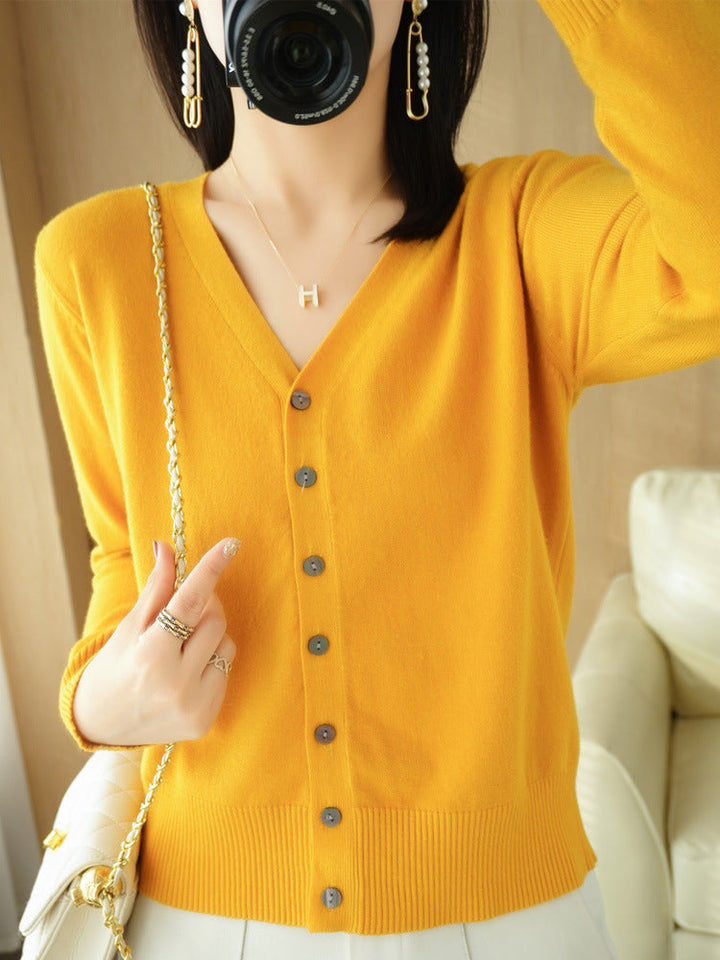 Femme portant un cardigan jaune doré à col en V, doté de boutons contrastés et d’une maille fine, associé à un sac à chaîne dorée et à un pantalon clair. Le style lumineux et moderne met en valeur une coupe élégante et féminine.