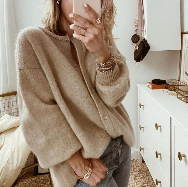 Une femme prend une photo devant un miroir, vêtue d'un cardigan marron oversize en maille douce boutonnée, associé à un jean décontracté et à plusieurs bracelets en or, dans une chambre lumineuse et élégamment décorée.