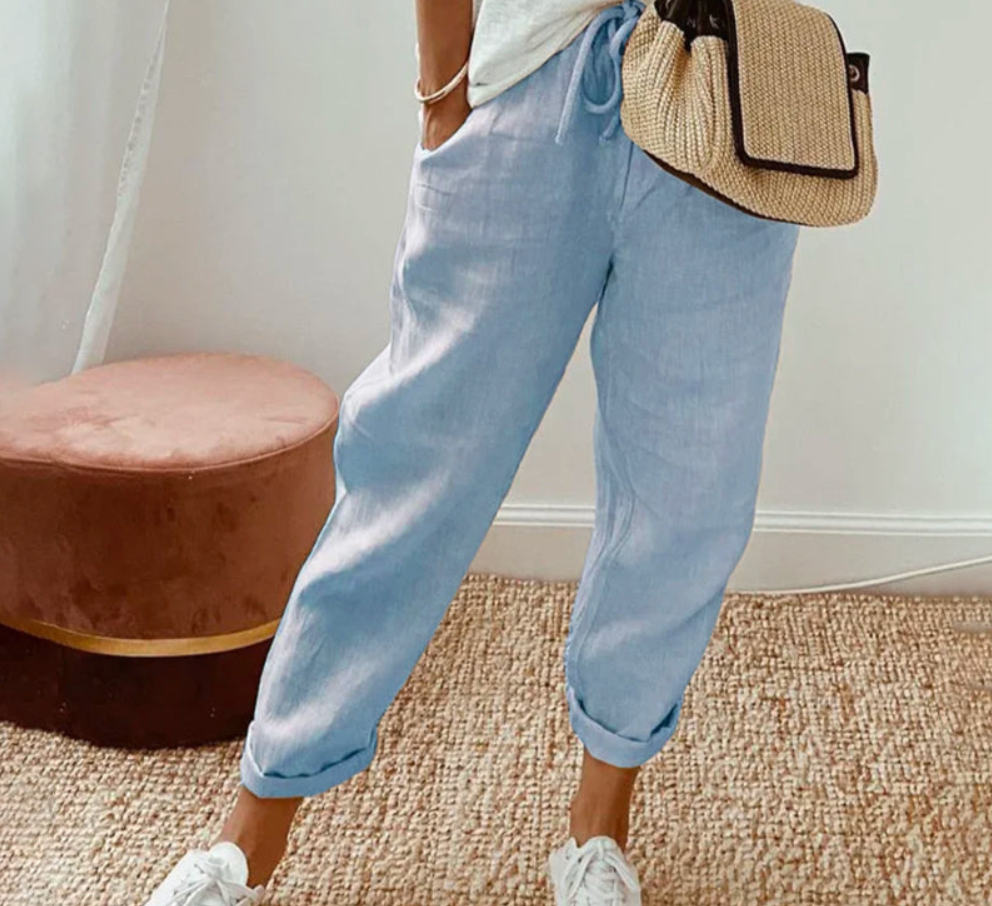 Pantalon ample pour femme en tissu léger bleu clair, coupe large et fluide avec taille élastiquée et cordon de serrage, porté de manière décontractée avec des baskets blanches et un sac en paille, idéal pour un style confortable et estival.