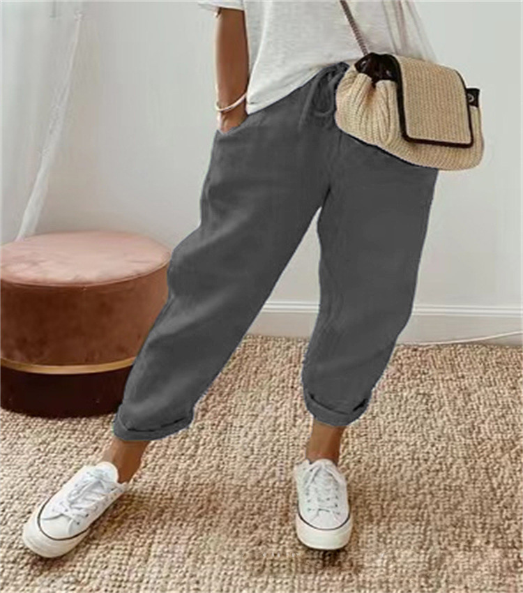 Pantalon ample pour femme en tissu léger gris foncé, à coupe large et fluide avec taille élastiquée et cordon de serrage, porté avec un t-shirt clair, des baskets blanches et un sac en paille pour un style décontracté et confortable.