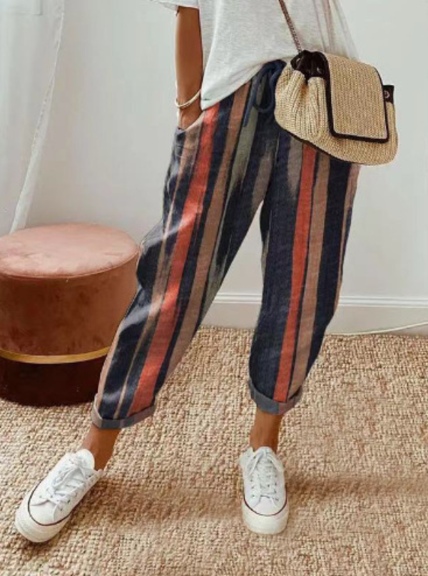 Pantalon ample pour femme en tissu léger à rayures verticales bleues, terracotta et beige, coupe large et fluide avec taille élastiquée et cordon de serrage, porté avec des baskets blanches et un sac en paille pour un style décontracté et moderne.