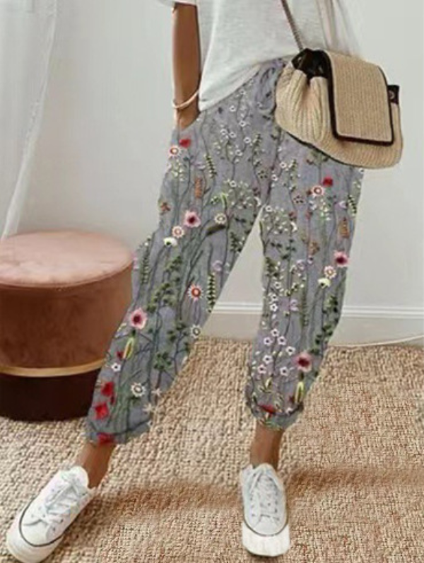 Pantalon ample pour femme en tissu léger gris à motif floral multicolore, coupe large et fluide avec taille élastiquée et cordon de serrage, porté avec un haut clair, des baskets blanches et un sac en paille pour un look décontracté et féminin.