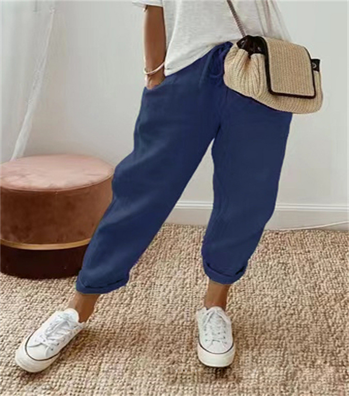 Pantalon ample pour femme en tissu léger bleu foncé, avec coupe large et fluide, taille élastiquée et cordon de serrage, porté avec un haut clair, des baskets blanches et un sac en paille pour un look décontracté et estival.