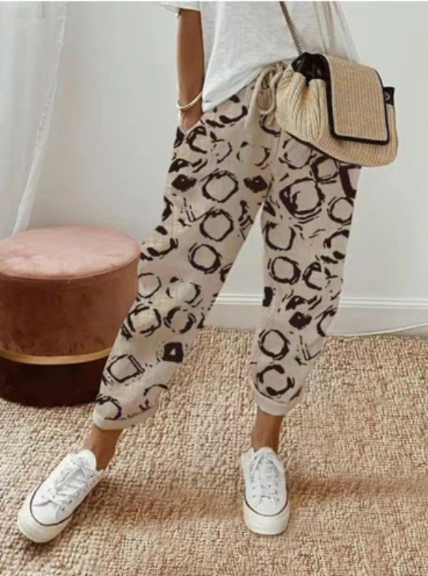 Pantalon ample pour femme en tissu léger beige à motifs abstraits marron, coupe large et fluide avec taille élastiquée et cordon de serrage, porté avec un haut clair, des baskets blanches et un sac en paille pour un look décontracté et tendance.