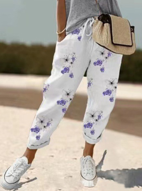 Pantalon ample pour femme en tissu léger blanc à motif floral violet et noir, coupe large et fluide avec taille élastiquée et cordon de serrage, porté avec un haut gris, des baskets blanches et un sac en paille pour un style estival et décontracté.