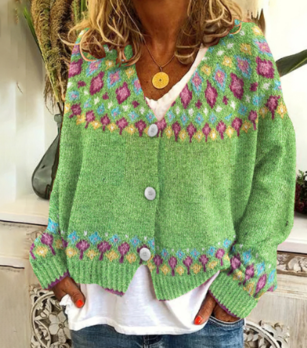Femme portant un cardigan en maille vert clair avec motifs géométriques colorés en rose, violet et bleu le long du col, des manches et de l’ourlet. Le cardigan est boutonné à l’avant et porté sur un t-shirt blanc ample et un jean décontracté. L’ensemble offre un style moderne, lumineux et bohème, idéal pour la mi-saison.