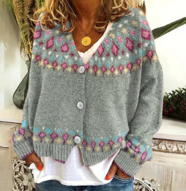 Une femme porte un cardigan gris en maille orné de motifs roses, violets et bleus sur le haut, les manches et le bas. Le cardigan se boutonne sur le devant et se porte par-dessus un t-shirt blanc ample et un jean décontracté. Ce look dégage une allure moderne, chaleureuse et bohème, idéale pour un style impeccable au quotidien.