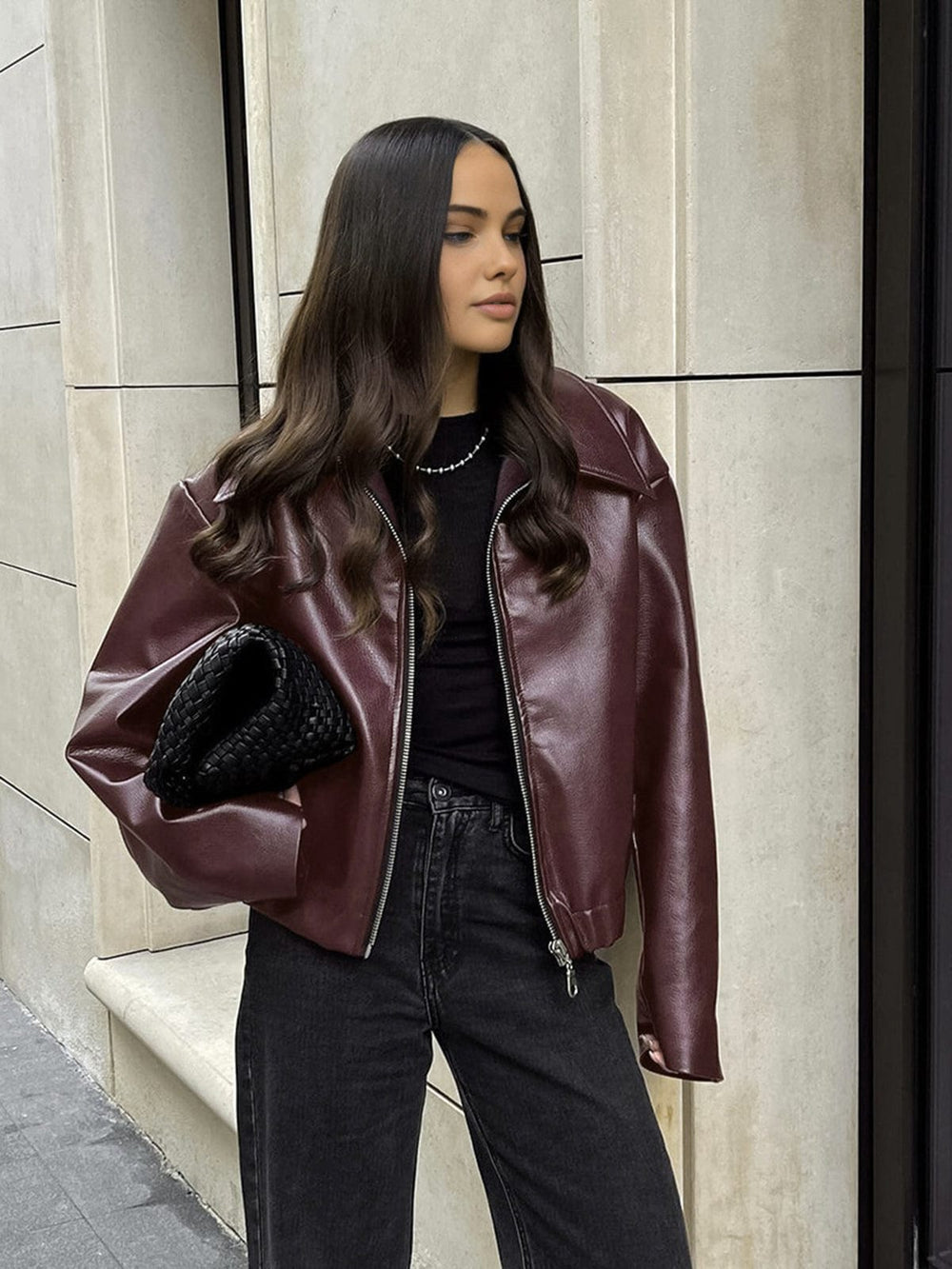 Femme portant une veste en simili cuir bordeaux, avec une fermeture zippée, un look moderne et élégant, assortie à un haut noir et un jean noir pour un style décontracté et chic.