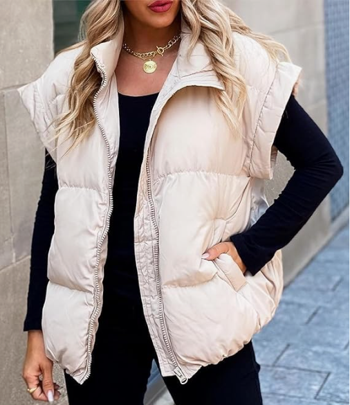 Une femme pose dans une rue pavée, portant un gilet matelassé sans manches de couleur beige clair. Le gilet est zippé à l’avant et légèrement oversize, offrant un style confortable et chaud pour la saison hivernale. Elle le porte par-dessus un haut noir à manches longues, assorti à un pantalon noir. Ses cheveux blonds ondulés tombent sur ses épaules, et elle complète la tenue avec un collier doré à pendentif, créant un look chic et moderne.