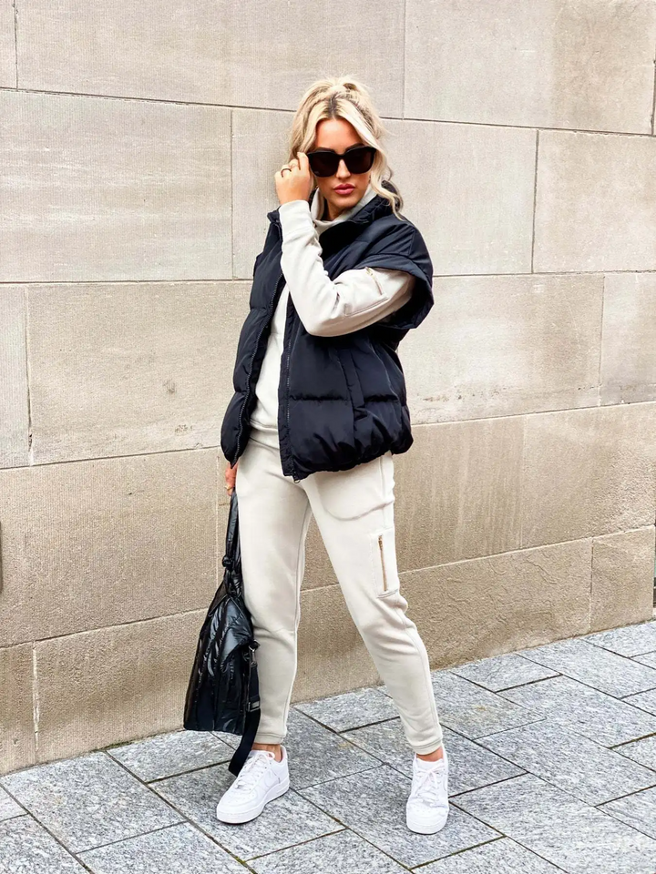 Une femme pose dans la rue, portant un gilet matelassé sans manches noir au style décontracté et moderne. Elle l’associe à un ensemble beige composé d’un sweat à manches longues et d’un pantalon de jogging assorti. Elle complète sa tenue avec des baskets blanches, un grand sac noir et des lunettes de soleil foncées. Ses cheveux blonds attachés en demi-queue ajoutent une touche chic à ce look d’hiver confortable et tendance.