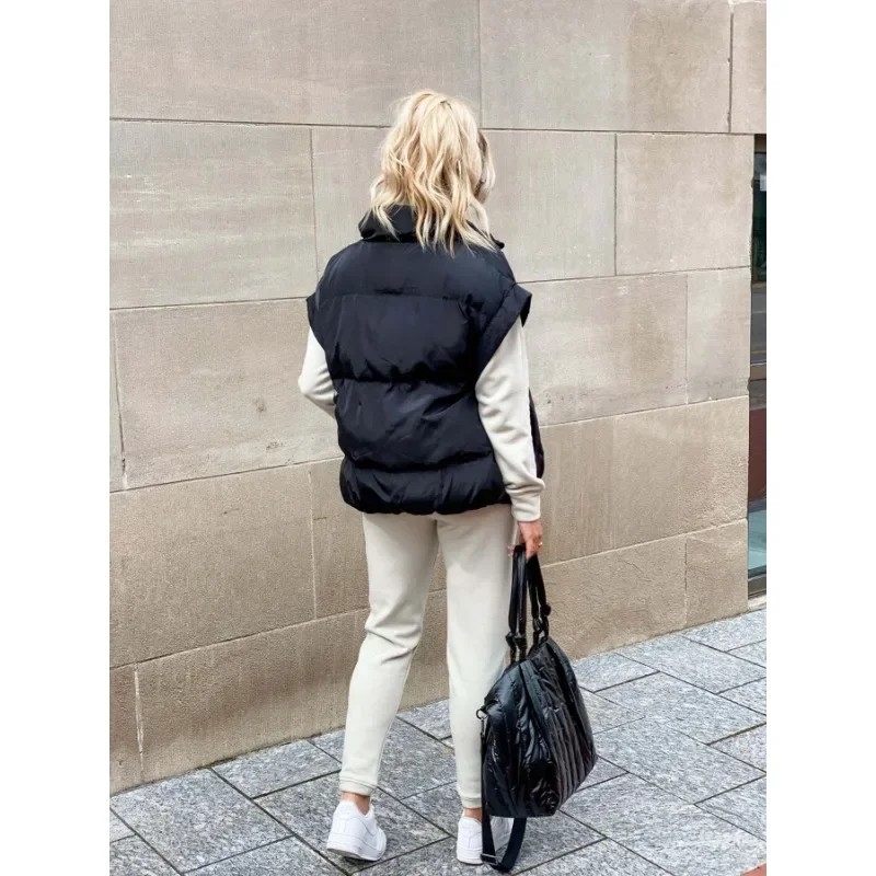 Vue arrière d’une femme marchant en extérieur, vêtue d’un gilet matelassé sans manches noir au style décontracté. Elle porte en dessous un ensemble beige composé d’un sweat à manches longues et d’un pantalon assorti. Ses cheveux blonds tombent librement sur ses épaules, et elle tient un grand sac noir à la main. La tenue, complétée par des baskets blanches, crée un look confortable et moderne idéal pour la saison froide.
