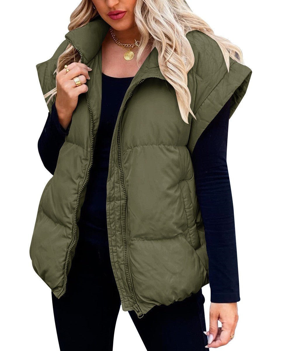 Une femme porte un gilet matelassé vert à fermeture éclair. Ce gilet à la coupe ample et moderne est idéal pour l'hiver. Elle le porte par-dessus un haut noir à manches longues et un pantalon noir, créant un contraste élégant. Ses cheveux blonds ondulés tombent librement sur ses épaules, et elle accessoirise sa tenue d'un collier en or et de plusieurs bagues pour un look à la fois chic et décontracté.