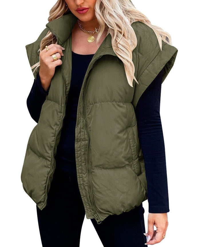 Une femme porte un gilet matelassé vert à fermeture éclair. Ce gilet à la coupe ample et moderne est idéal pour l'hiver. Elle le porte par-dessus un haut noir à manches longues et un pantalon noir, créant un contraste élégant. Ses cheveux blonds ondulés tombent librement sur ses épaules, et elle accessoirise sa tenue d'un collier en or et de plusieurs bagues pour un look à la fois chic et décontracté.