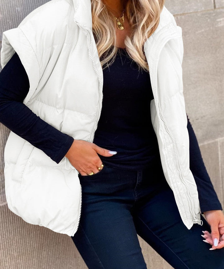 Une femme assise contre un mur porte un gilet matelassé sans manches blanc à fermeture éclair sur le devant. Le gilet présente une coupe ample et confortable, parfaite pour les journées froides. Elle le combine avec un haut noir à manches longues et un pantalon noir ajusté. Ses cheveux blonds bouclés encadrent son visage, et elle accessoirise sa tenue avec un collier doré et des bagues, créant un style élégant et moderne pour l’hiver.