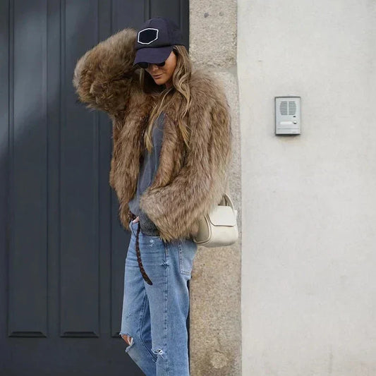 Nicole | Femme portant une veste longue en fourrure marron, avec un t-shirt gris, un jean déchiré et un sac à main blanc, ajustant sa casquette noire tout en souriant, devant une porte noire, en mode décontracté pendant l'hiver.