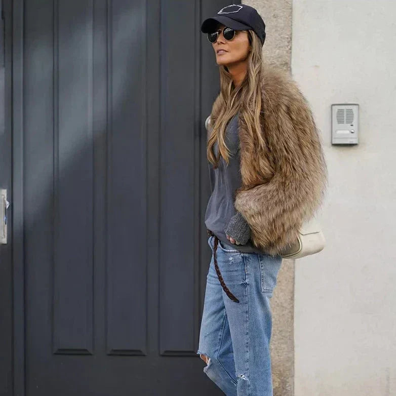Nicole | Femme portant une veste longue en fourrure marron, avec un t-shirt gris et un sac à main blanc, souriant tout en se tenant devant une porte noire, avec des lunettes de soleil et une casquette noire, en mode décontracté pendant l'hiver.