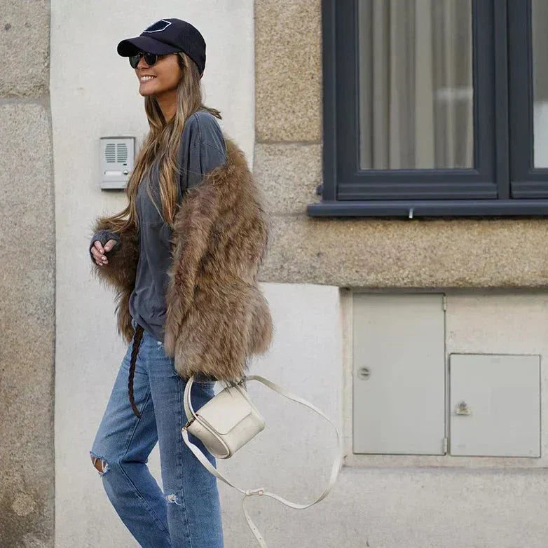 Nicole | Veste longue en fourrure marron portée par une femme souriante, vêtue d'un t-shirt gris et de jeans déchirés, avec un sac à main blanc, se promenant dans la rue par une journée ensoleillée.