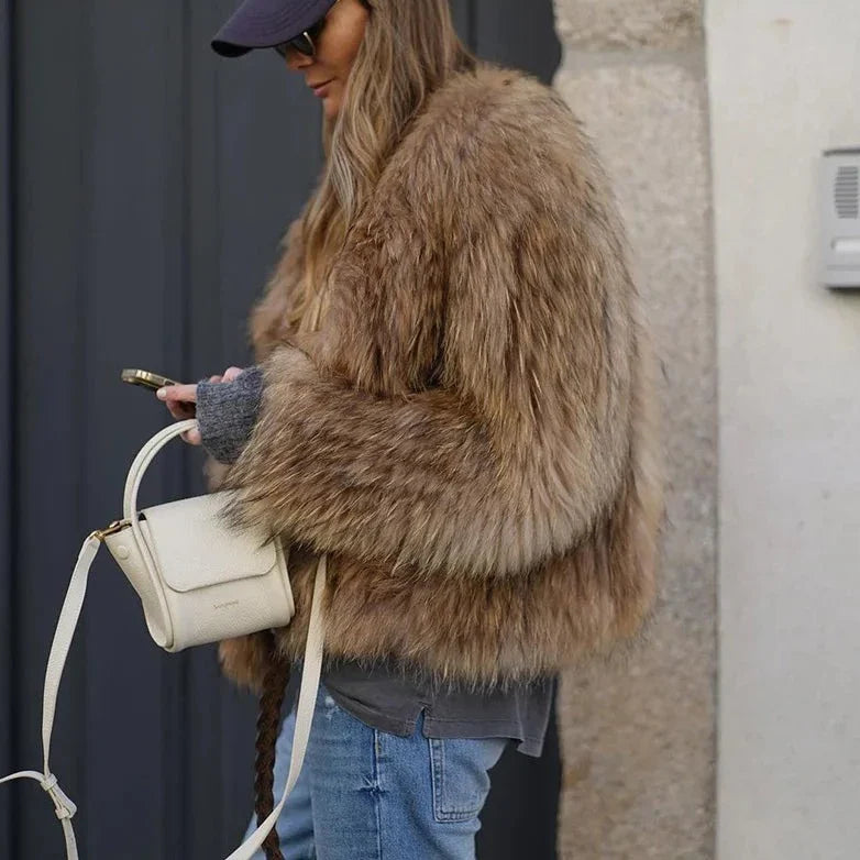 Nicole | Veste longue en fourrure marron portée par une femme avec une petite sacoche blanche et un look décontracté, tout en consultant son téléphone portable, idéale pour l'hiver.