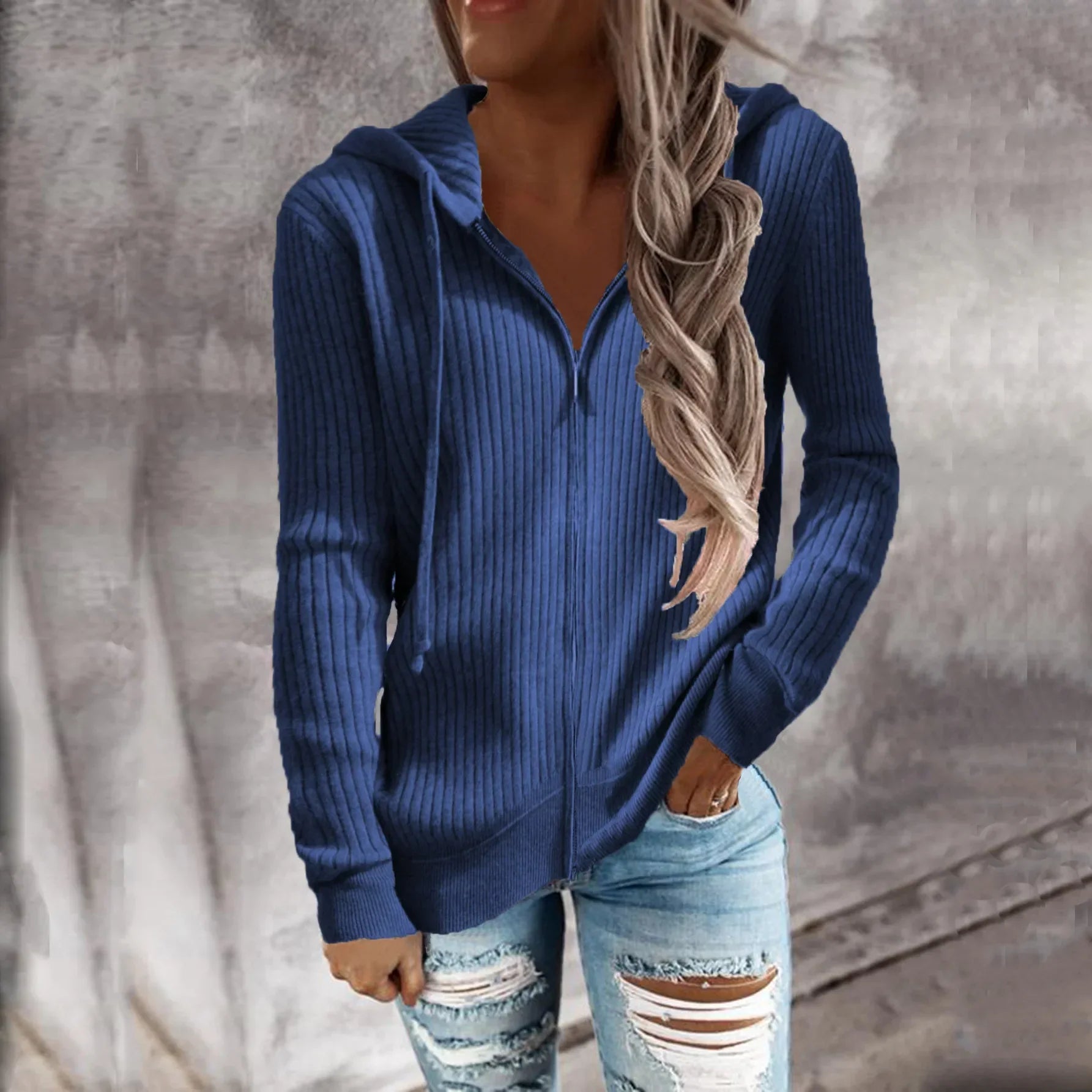 Veste à capuche zippée en maille côtelée, coloris bleu, portée par une femme souriante. Elle porte un jean déchiré et une tenue décontractée, idéale pour les journées fraîches d'automne et d'hiver.