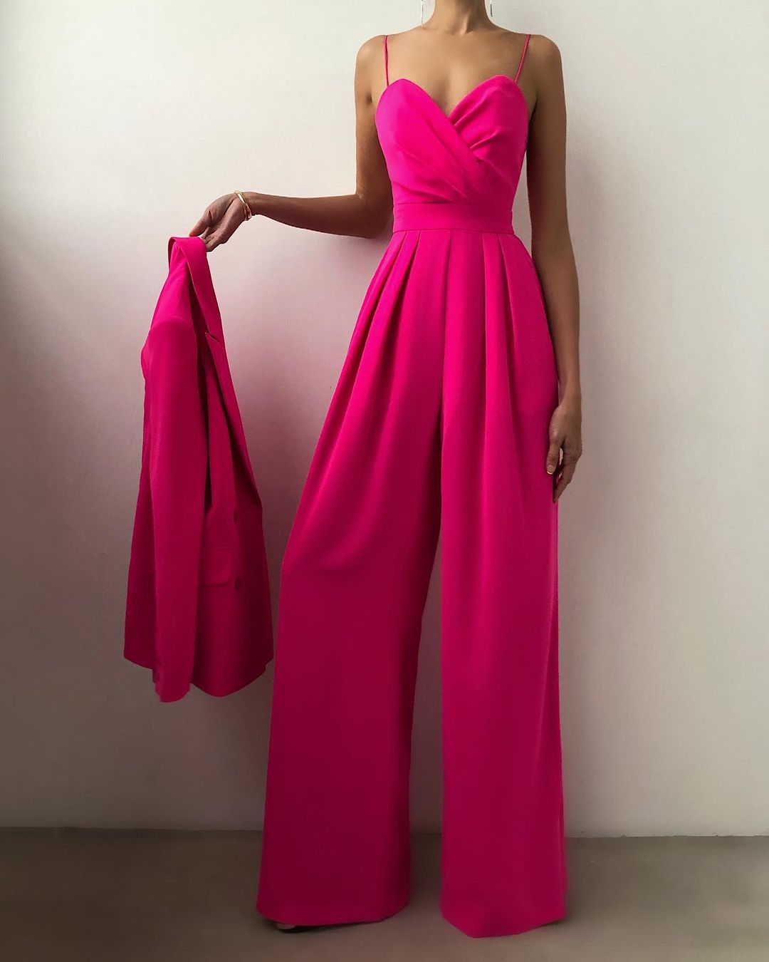 Combinaison longue fuchsia à fines bretelles, décolleté cœur croisé et taille ceinturée, avec pantalon large fluide, portée par un mannequin tenant une veste assortie, sur fond neutre.