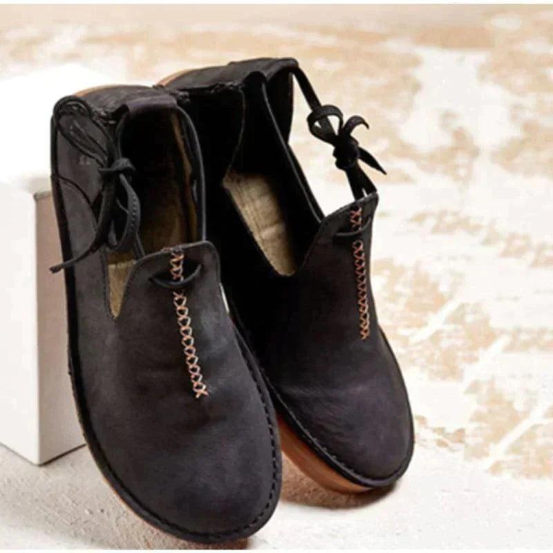Chaussures d’automne pour femme en cuir noir, au style moderne et minimaliste, avec coutures contrastées apparentes, lien décoratif sur le côté, ouverture souple et semelle plate confortable, présentées sur un fond clair.