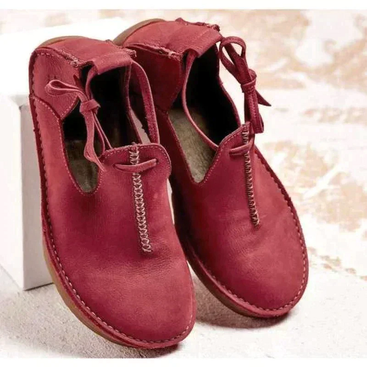 Chaussures d’automne pour femme en cuir rouge bordeaux, au style moderne et épuré, avec coutures apparentes faites main, lien décoratif sur le côté, ouverture souple et semelle plate confortable, présentées sur un fond clair.